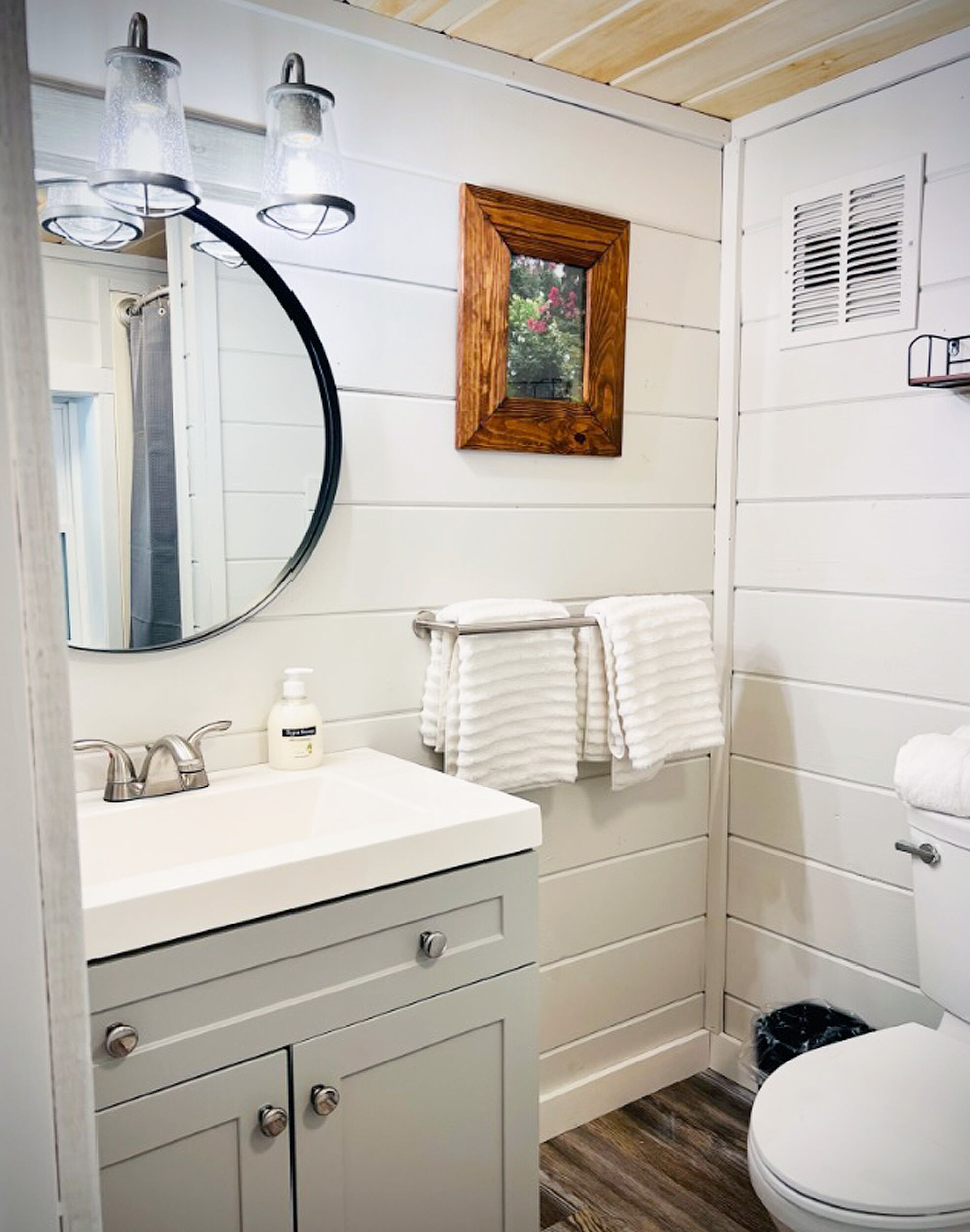 Une salle de bain avec un meuble-lavabo blanc, un miroir rond, une fenêtre à cadre en bois et des toilettes.