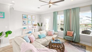 Ein Wohnzimmer mit einem beigefarbenen Ecksofa, einem runden Couchtisch und einem Deckenventilator. Zwei Fenster mit grünen Vorhängen, eine Tür und eine Wand mit gerahmten Bildern sind zu sehen.