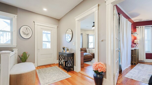 A spacious hallway with hardwood flooring, a white bench, a small table with a vase of flowers, and a door leading to a living area. - Old Brookline Charm 5 minLongwood In-Unit Laundry (Brookline)