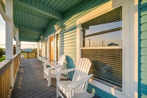 Ein überdachter Balkon mit Holzgeländern, zwei Adirondack-Stühlen und Blick auf ein Haus durch ein Fenster mit Jalousien.
