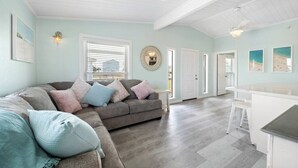 Ein geräumiges Wohnzimmer mit einem grauen Ecksofa, einem kleinen weißen Tisch und einer Küchenzeile mit weißer Arbeitsplatte und Hockern.
