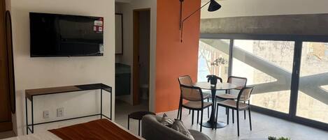 A modern hotel room with a bed, a sofa, a dining area with chairs, a TV mounted on the wall, and large windows offering a view.
