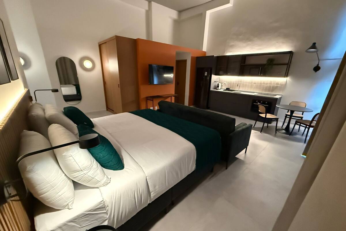 A modern hotel room with a large bed, a sofa, a desk, and a kitchenette.