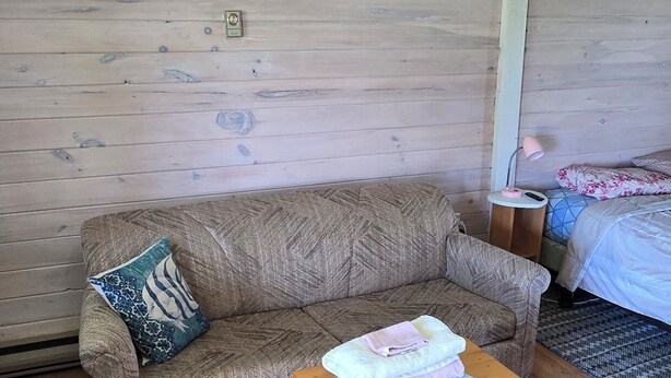 A room with a wooden floor, a sofa with a patterned cushion, a small wooden table with towels, and a bed with a patterned bedspread. - Suite 9 - Older/Simple/Rustic/No Frills/Budget 1 Bed - Near Cabot Beach Park (Kensington)