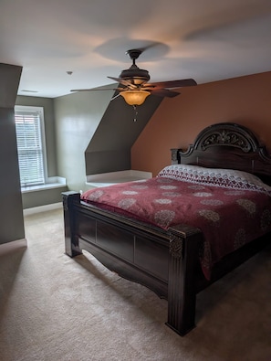 A bedroom with a sloped ceiling, a ceiling fan, a bed with a patterned bedspread, and a window with blinds. - Renewal Room at the Hilltop Hideaway King Bed (Hampstead)