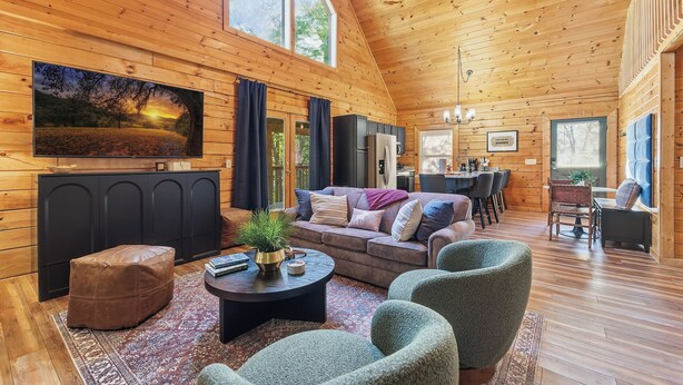 Ein gemütliches Blockhaus-Interieur mit Holzbalkendecke, einem großen an der Wand montierten Fernseher, einer Couch und einem Essbereich.