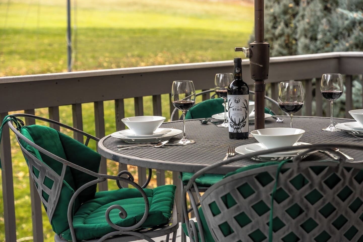 A round metal table with white plates, bowls, and wine glasses, set on a balcony with green cushioned chairs.