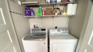 Un cuarto de lavandería con lavadora y secadora de carga frontal, un estante superior con productos de limpieza y una puerta a la izquierda.
