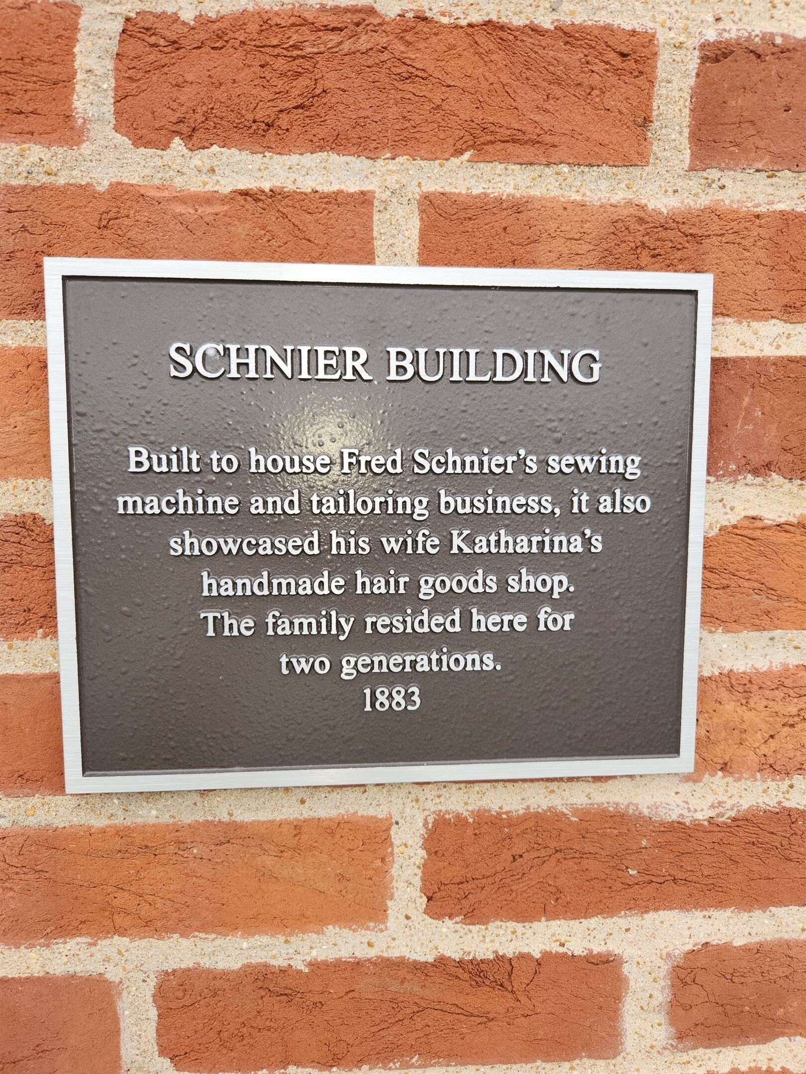 Een plaquette op een bakstenen muur met de tekst 'SCHNIER BUILDING' en historische informatie over het bedrijf en woonhuis van Fred Schnier.
