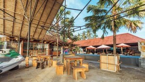 An outdoor seating area with wooden tables and chairs, a covered structure with a thatched roof, and palm trees in the background. - Semaya One Beach Hotel (Bali)