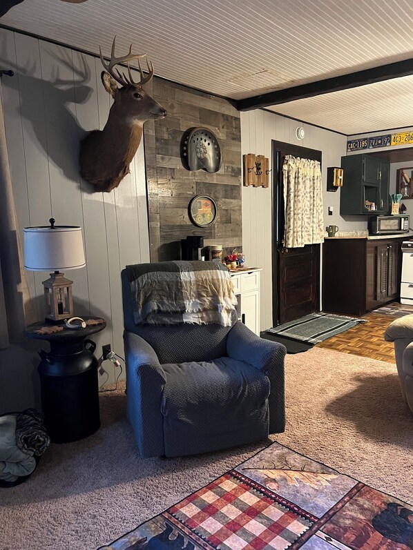 A living room with a mounted deer head, a fireplace, a cozy armchair, and a kitchen area in the background.