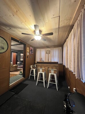 A bar area with wooden walls, a ceiling fan, and three white bar stools.