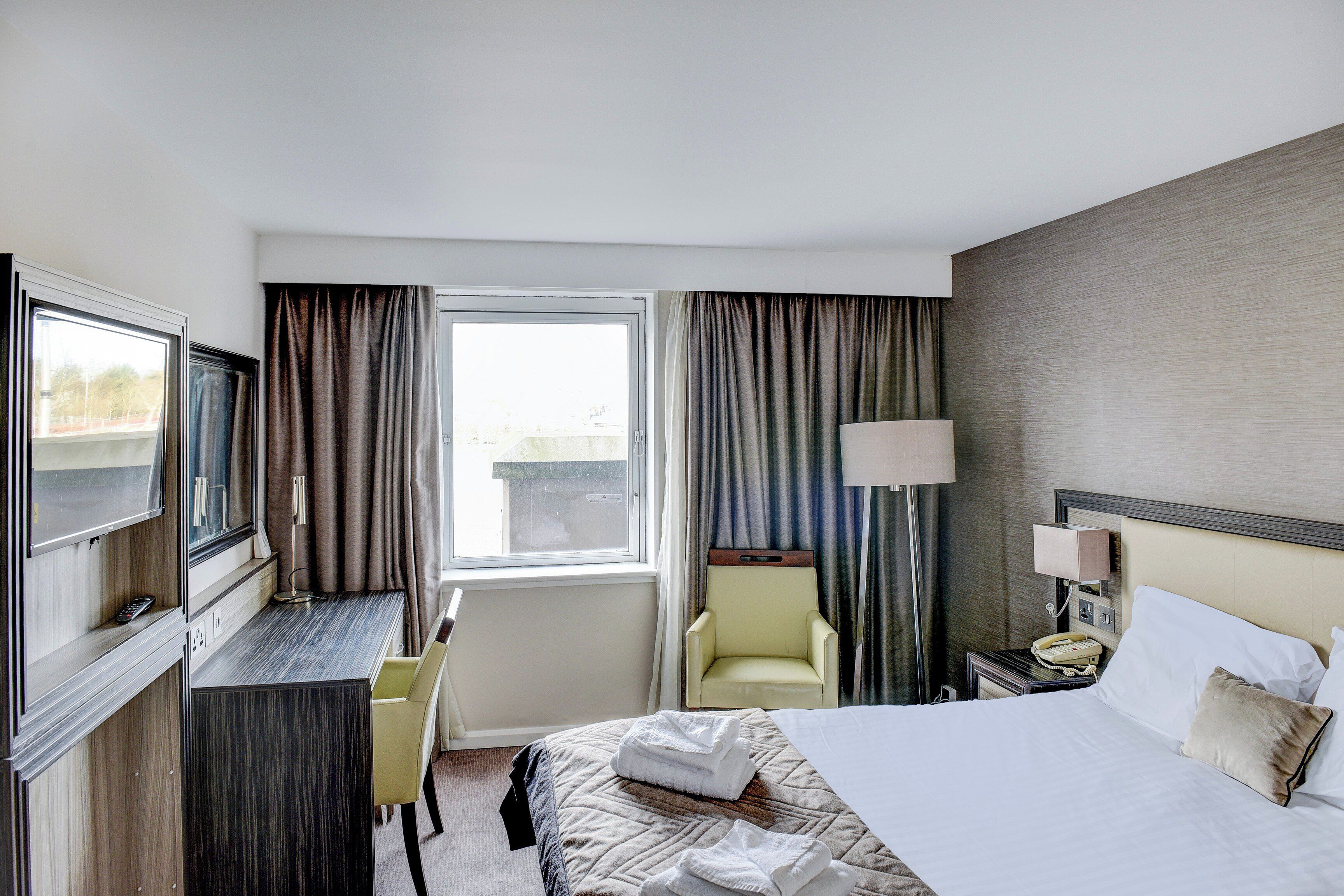 A hotel room with a bed, a desk, a chair, a TV, a lamp, and a window with curtains.