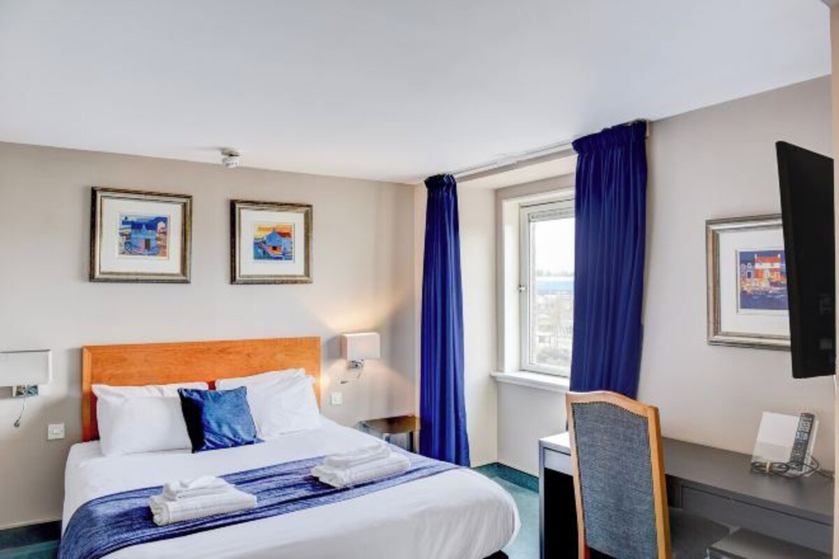 A hotel room with a bed, a desk, a chair, a window with curtains, and framed pictures on the wall.