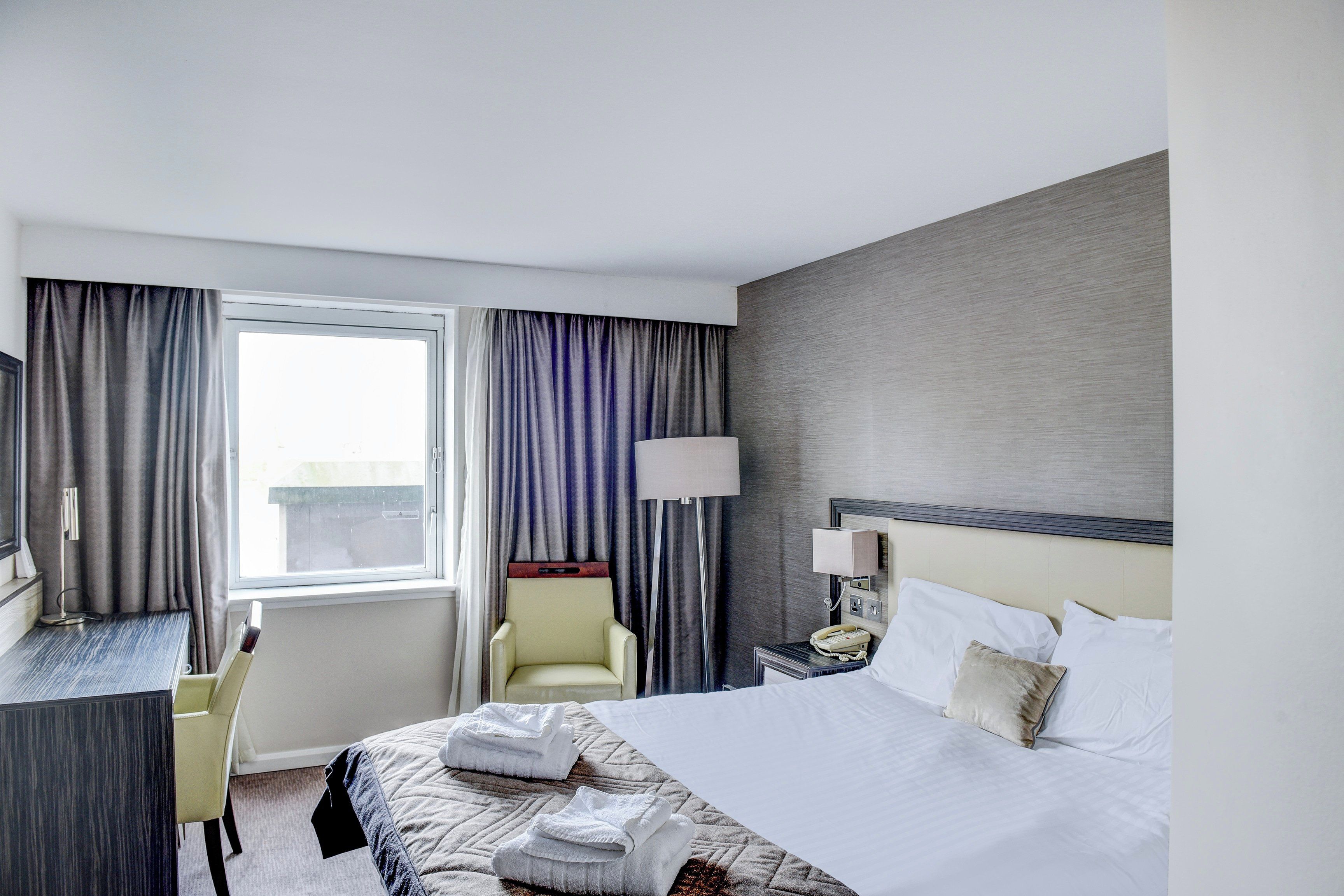 Habitación de hotel con cama, escritorio, silla, lámpara y ventana con cortinas.