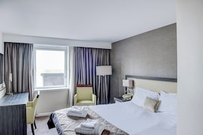 Une chambre d’hôtel avec un lit, un bureau, une chaise, une lampe et une fenêtre avec des rideaux.