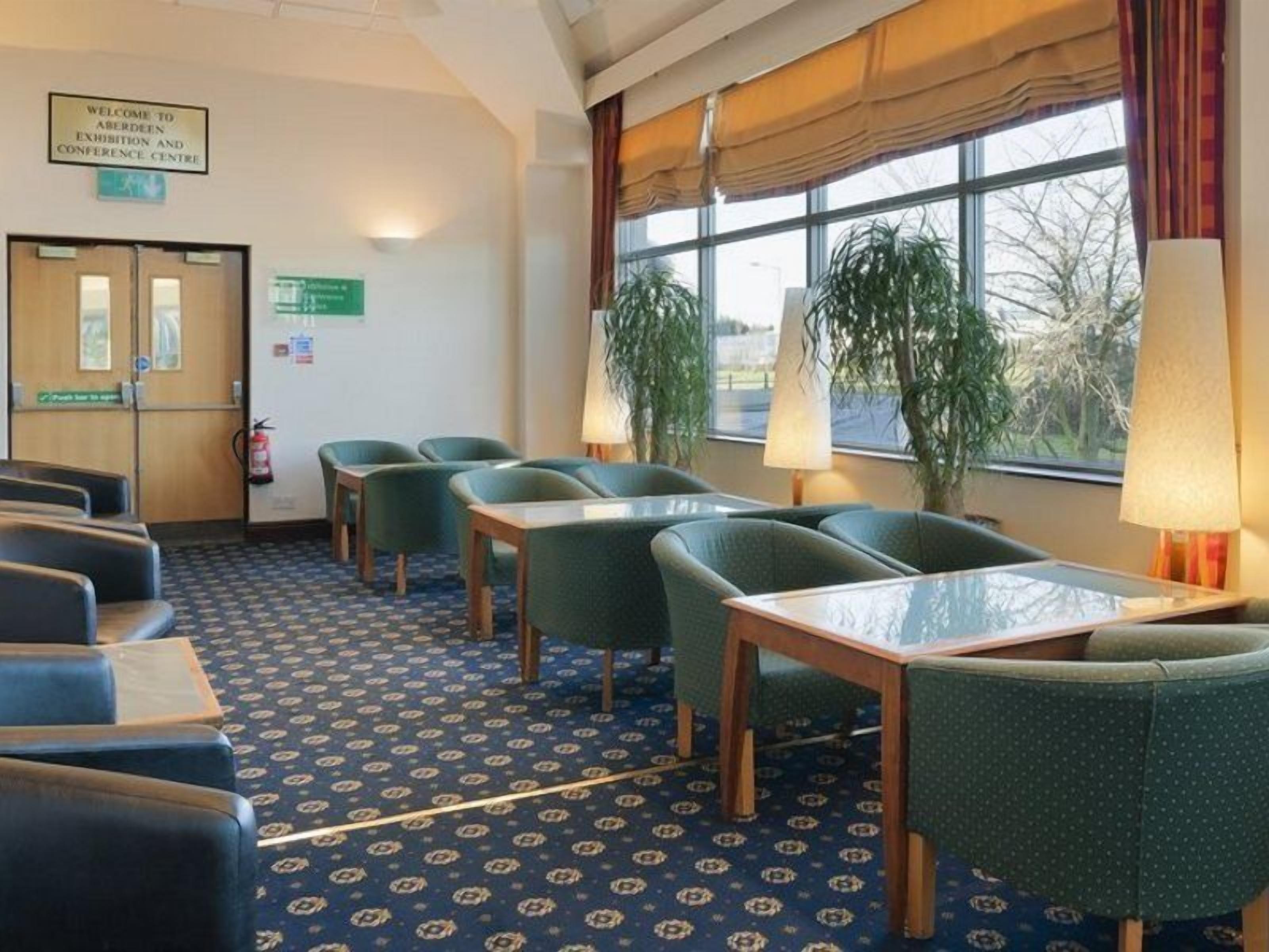Un salón de conferencias de hotel con alfombra estampada en azul, sillas tapizadas en verde, una mesa de madera y ventanas amplias con cortinas.