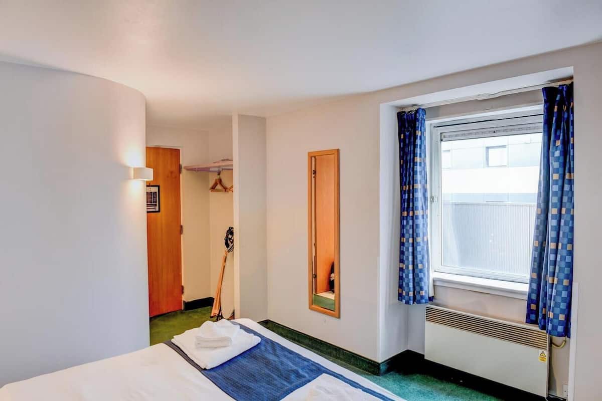 Una habitación de hotel con cama, ventana con cortinas, espejo y perchero.