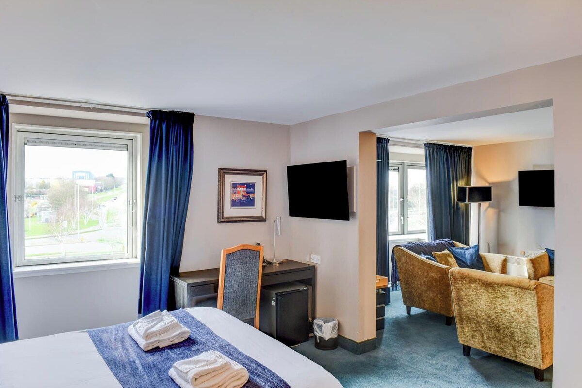 Habitación de hotel con cama, escritorio, silla, sofá, televisión y ventana con cortinas.