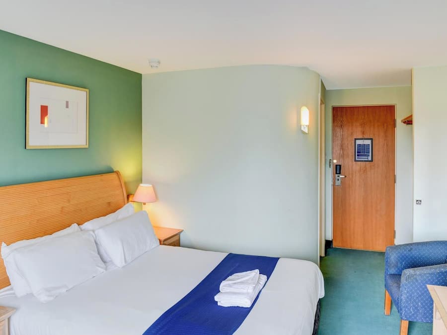 A hotel room with a bed, a nightstand, a lamp, a door, and a chair.