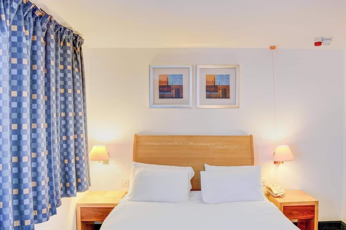 Habitación de hotel con cama, dos lámparas de noche, mesita con teléfono y ventana con cortinas azules.