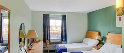 A hotel room with two beds, a desk with lamps, a chair, and a window with curtains.