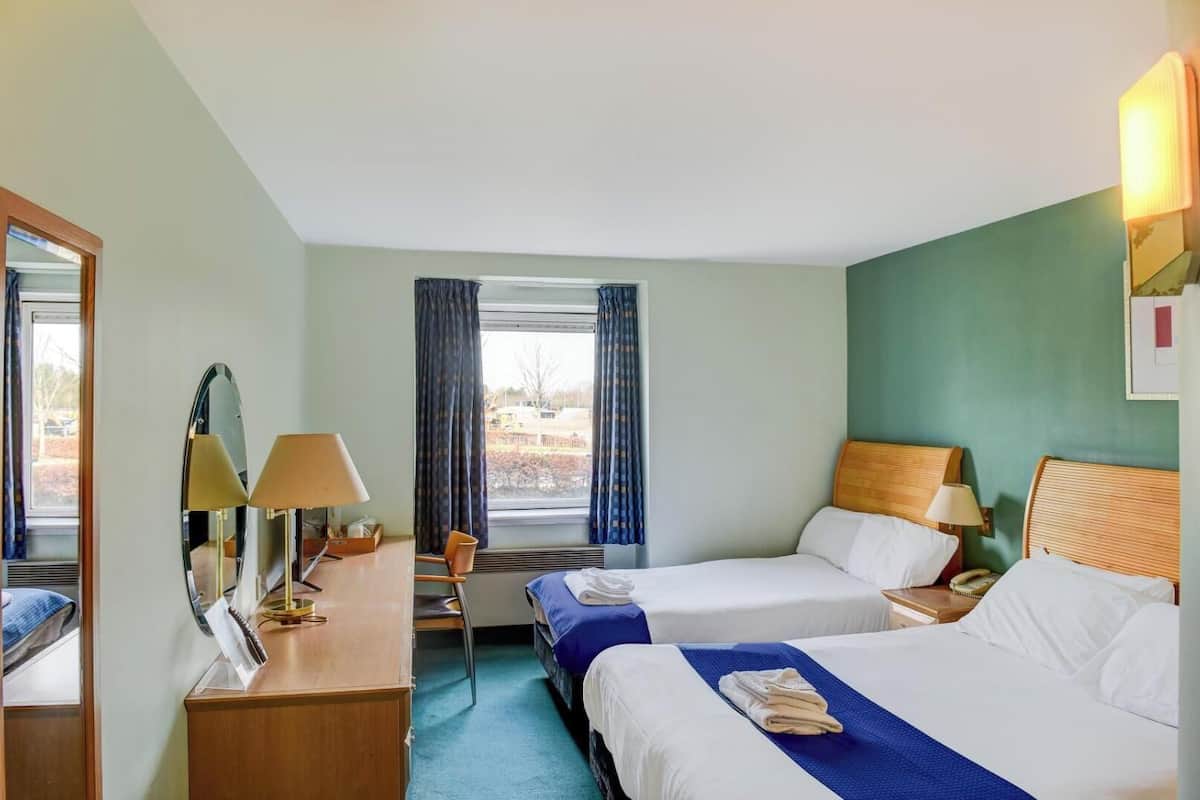 Habitación de hotel con dos camas, un escritorio con lámparas, una silla y una ventana con cortinas.