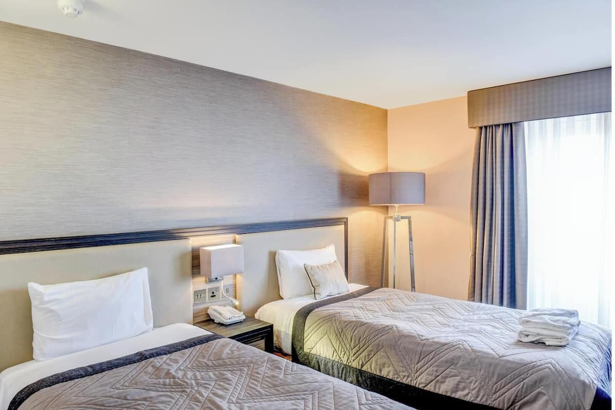 Habitación de hotel con dos camas, una mesita de noche con lámpara, teléfono y una ventana con cortinas.