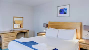 Habitación de hotel con una cama, cabecera de madera, mesita de noche con lámpara, escritorio con espejo y un cuadro abstracto enmarcado en la pared.