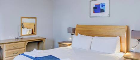 Habitación de hotel con una cama, cabecera de madera, mesita de noche con lámpara, escritorio con espejo y un cuadro abstracto enmarcado en la pared.