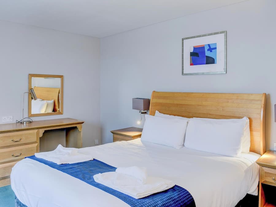 Habitación de hotel con una cama, cabecera de madera, mesita de noche con lámpara, escritorio con espejo y un cuadro abstracto enmarcado en la pared.