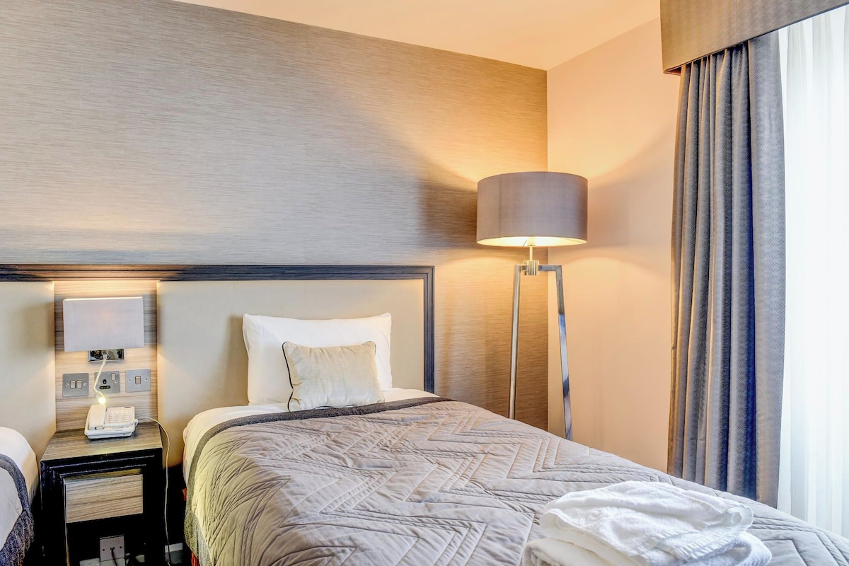 Una habitación de hotel con cama, lámparas de noche, un teléfono y una ventana con cortinas.