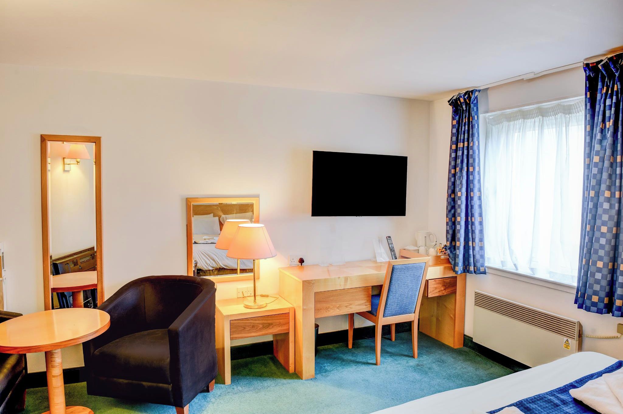 Une chambre d’hôtel comprenant un lit, un bureau avec une chaise, une petite table, une lampe, un téléviseur et une fenêtre avec des rideaux.