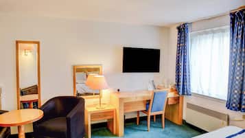 Une chambre d’hôtel comprenant un lit, un bureau avec une chaise, une petite table, une lampe, un téléviseur et une fenêtre avec des rideaux.