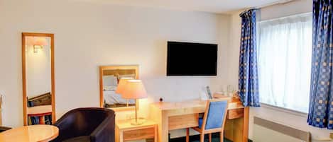 A hotel room with a bed, a desk with a chair, a small table, a lamp, a TV, and a window with curtains.