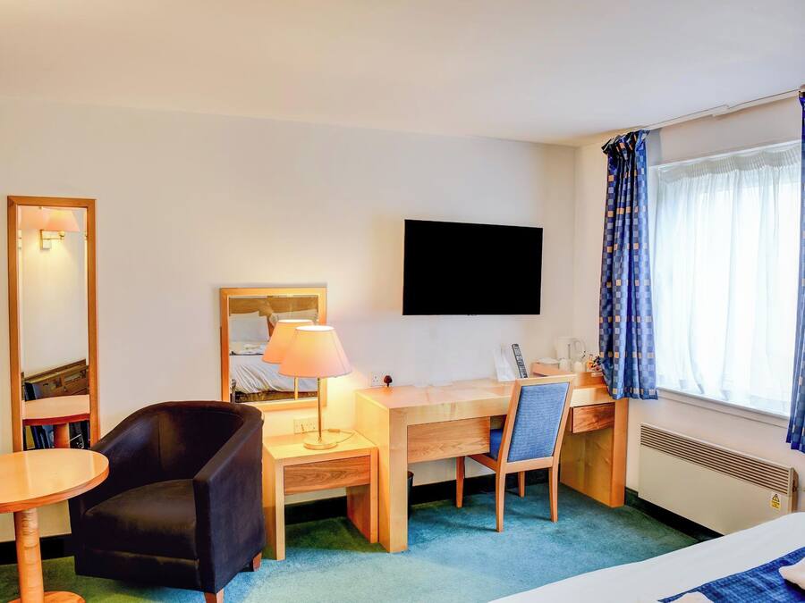 A hotel room with a bed, a desk with a chair, a small table, a lamp, a TV, and a window with curtains.