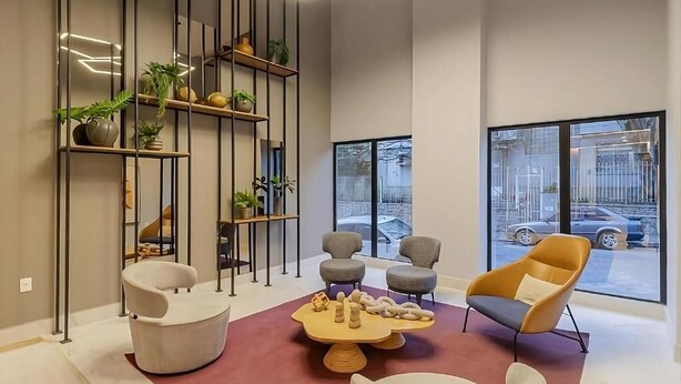 Un intérieur moderne comprenant un coin salon, une grande étagère avec des plantes et de grandes fenêtres donnant sur la rue.