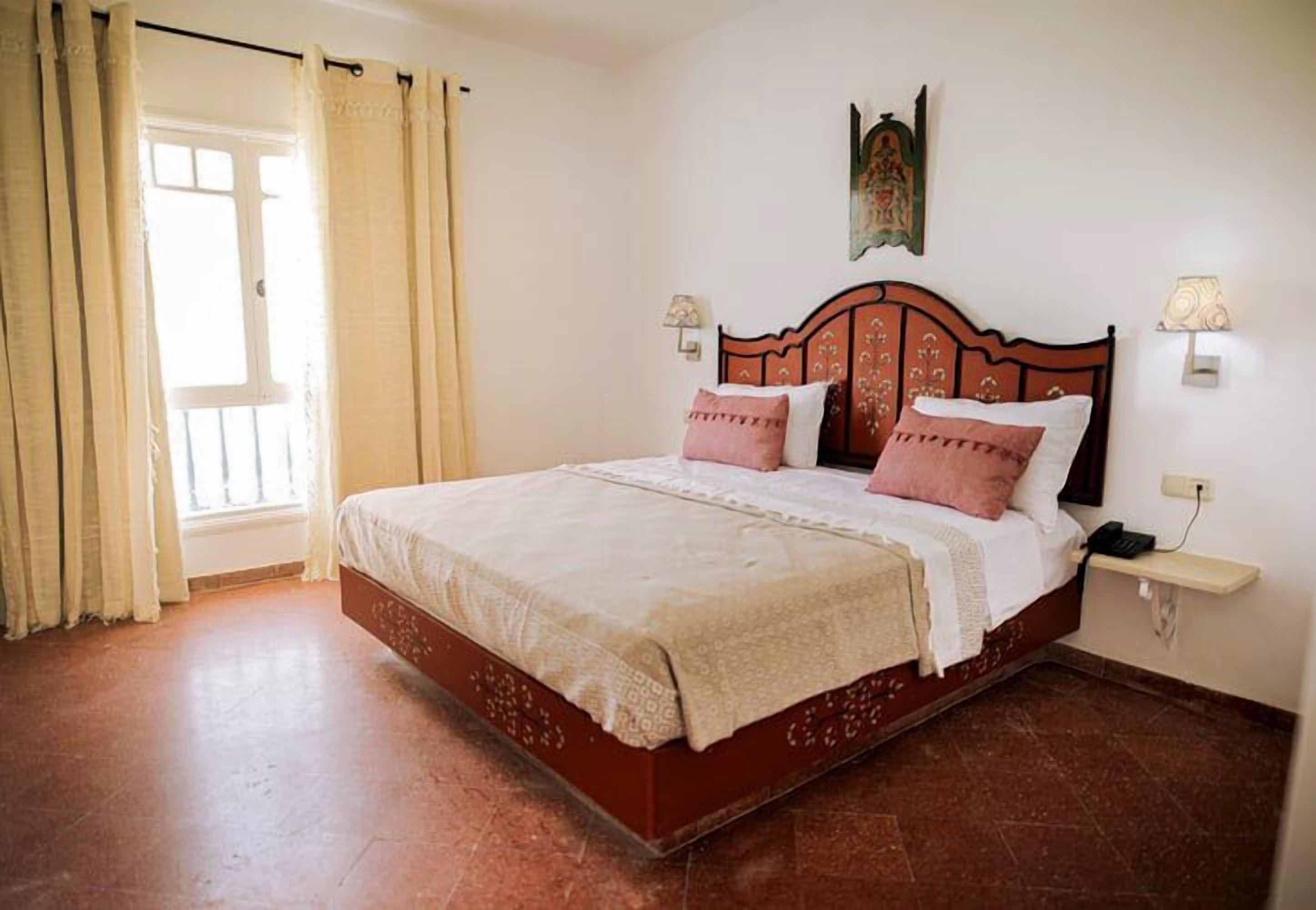 A bedroom with a wooden bed, beige curtains, a wall-mounted lamp, and a small shelf.