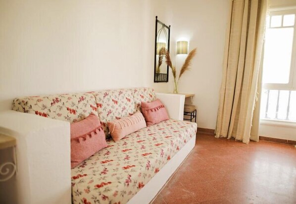 A living room with a floral sofa, a small side table, a lamp, and a window with curtains.