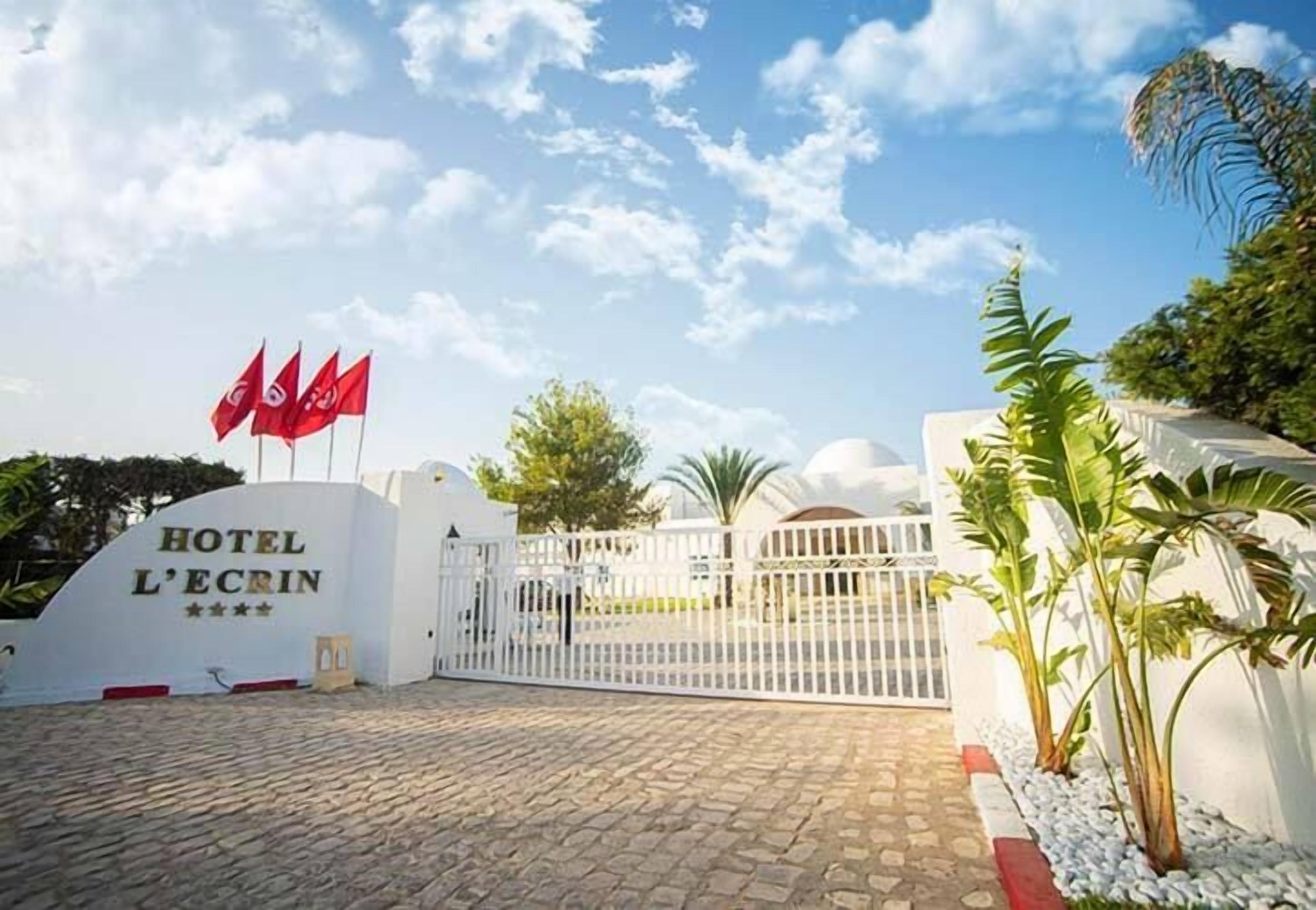 A hotel entrance with a white gate, flags, and a sign reading "HOTEL L'ECRIN".