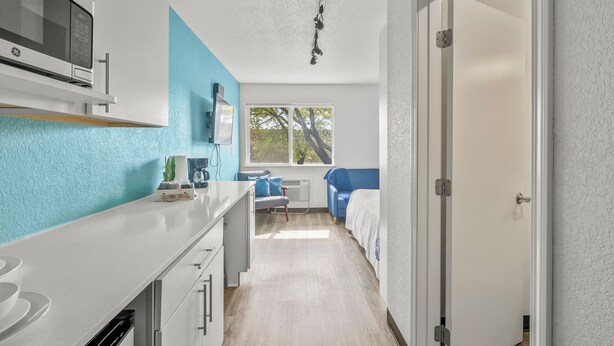 A compact kitchen with a microwave, a sink, and a window providing natural light. A bed with a blue headboard is visible in the adjacent room.