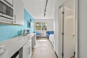 Eine kleine Küche mit Mikrowelle, Spüle und einem Fenster, das für natürliches Licht sorgt. Im angrenzenden Raum ist ein Bett mit blauer Kopfteil zu sehen.