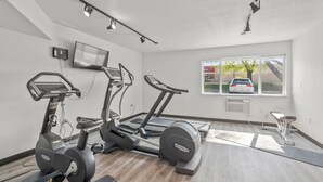 A modern gym with treadmills, a stationary bike, and a rowing machine. There is a TV mounted on the wall and a window with a view of a car and trees.