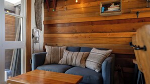 A cozy room with a wooden wall, a blue sofa with striped pillows, a wooden table, and a shelf with books and a Rubik's cube. - Rustic Mokau Escape with Ocean Views & Sauna (Mokau)