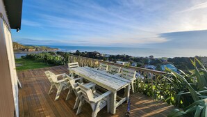 A wooden deck with a table and chairs, overlooking a coastal view with houses and greenery. - Rustic Mokau Escape with Ocean Views & Sauna (Mokau)