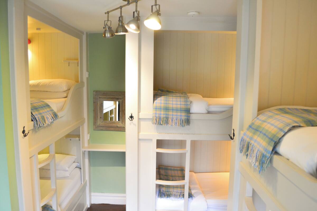 A bunk bed room with three levels, each with a pillow and a blanket. There are shelves below the beds and a mirror on the wall.
