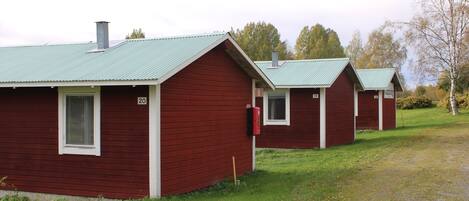 A row of red cabins with green roofs, numbered 19 and 20, set in a grassy area with trees in the background.