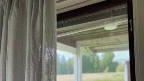 A view through a window with a white curtain, a sliding door, and a glimpse of a green lawn and a chair outside. - Borgsjö Strand (Erikslund)