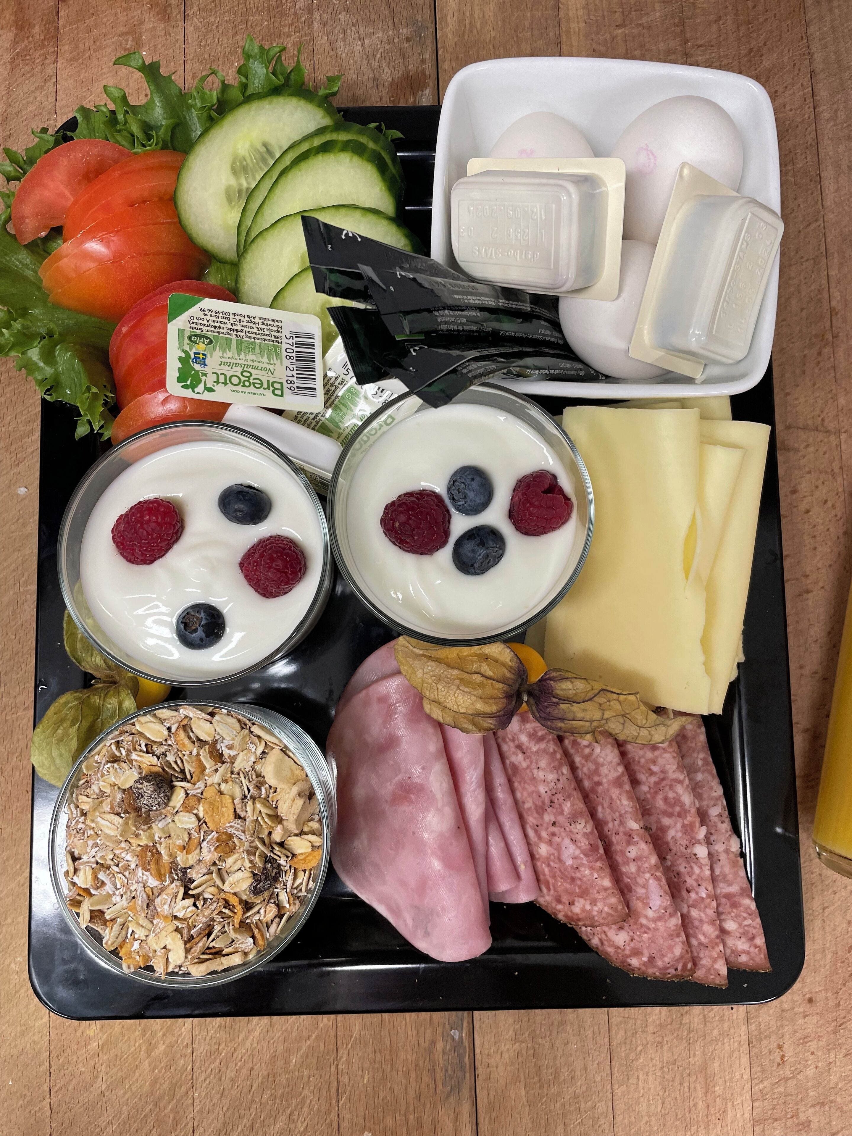 A tray with various food items including yogurt with berries, cheese, ham, salami, vegetables, and a container of granola.