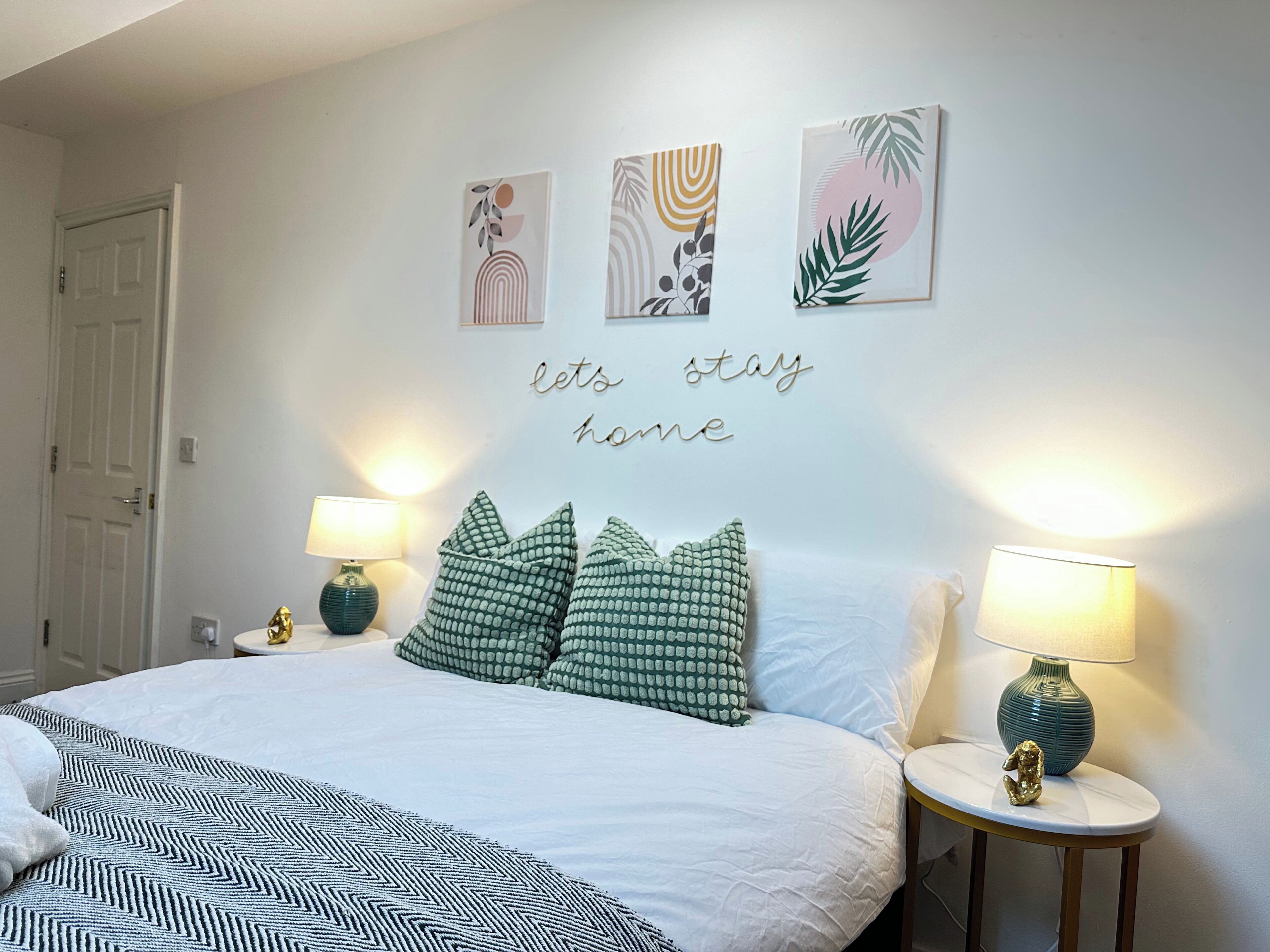 A bedroom with a bed, pillows, two bedside lamps, a wall with artwork, and a door.